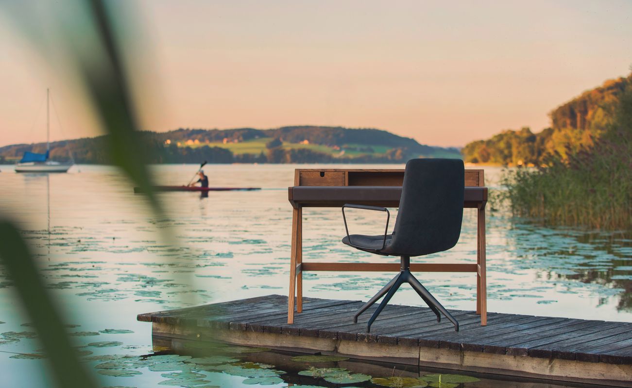 Ein moderner Schreibtischstuhl steht auf einem Holzsteg am Wasser, im Hintergrund sind Boote und eine ruhige Landschaft zu sehen.