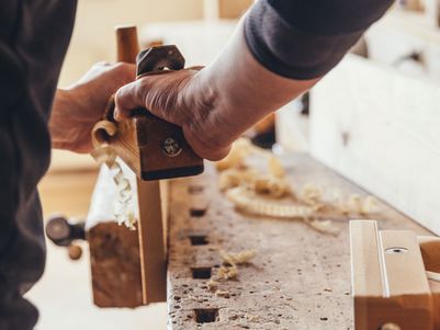 Ein Handwerker benutzt eine Hobelmaschine, um Holz zu bearbeiten. Späne liegen auf der Werkbank.