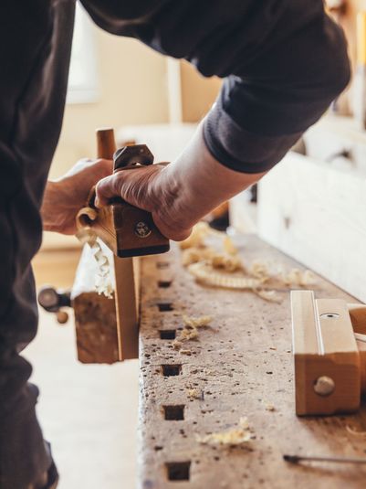 Ein Handwerker benutzt eine Hobelmaschine, um Holz zu bearbeiten. Späne liegen auf der Werkbank.