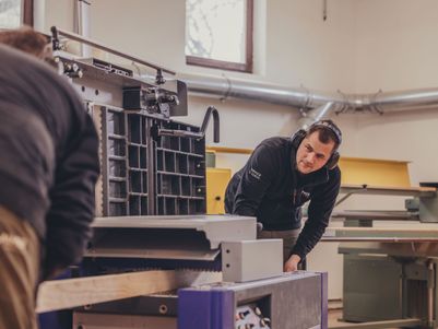 Zwei Personen in einer Werkstatt, eine Person bedient eine Maschine, während die andere Holz bearbeitet. Beide tragen Schutzkopfhörer.