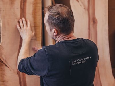 Ein Mann betrachtet eine Holzplatte in einer Werkstatt, mit Holzstücken im Hintergrund. Er trägt ein schwarzes Sweatshirt mit Aufdruck.