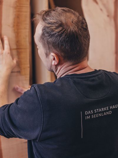 Ein Mann betrachtet eine Holzplatte in einer Werkstatt, mit Holzstücken im Hintergrund. Er trägt ein schwarzes Sweatshirt mit Aufdruck.