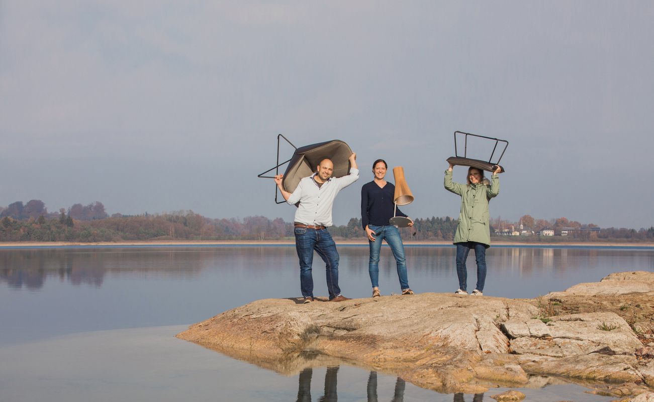 Drei Personen stehen auf einem Felsen am Wasser und halten Tische über ihren Köpfen. Die Landschaft ist ruhig mit einem klaren Himmel.