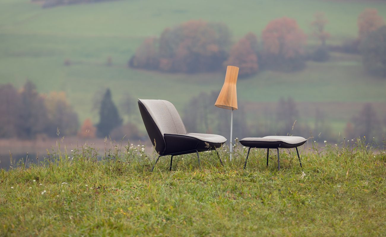 Zwei moderne Stühle und ein Tisch mit einer Lampe stehen auf einer Wiese mit Blick auf eine grüne Landschaft und Bäume im Hintergrund.