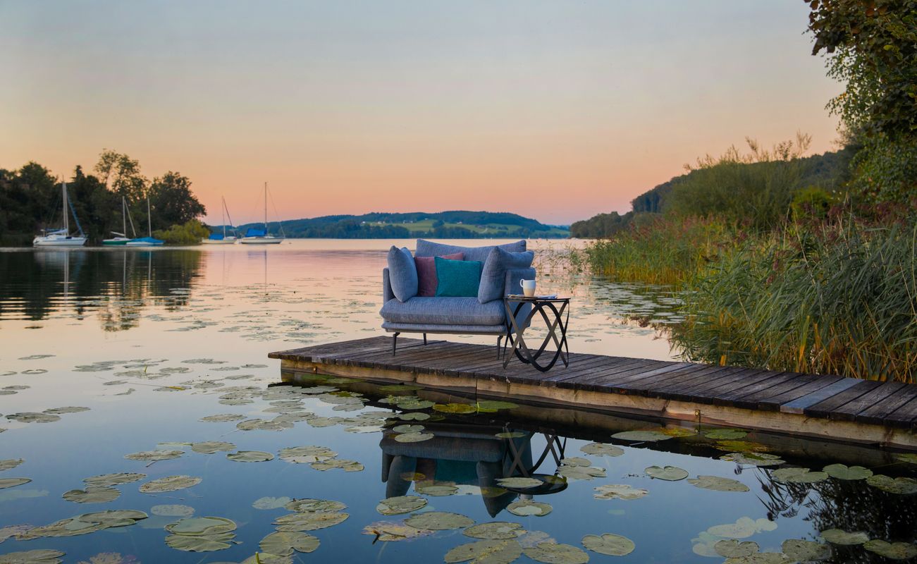 Eine gemütliche blaue Couch mit Kissen und ein kleiner Tisch auf einem Holzsteg am Wasser, umgeben von Seerosen und sanften Hügeln im Hintergrund.