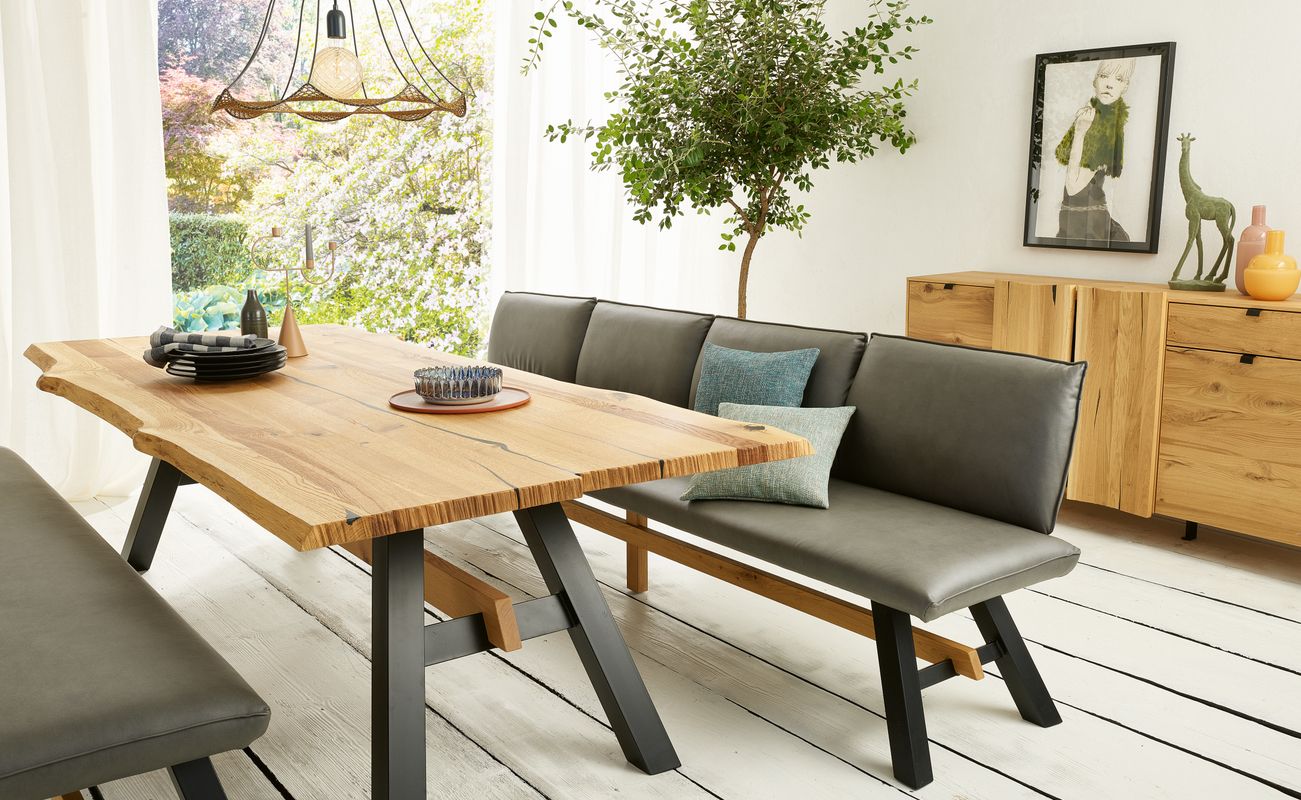 Ein moderner Essbereich mit einem Holztisch, grauen Bänken, einer Pflanze und einem Sideboard. Helle Wände und große Fenster sorgen für Licht.