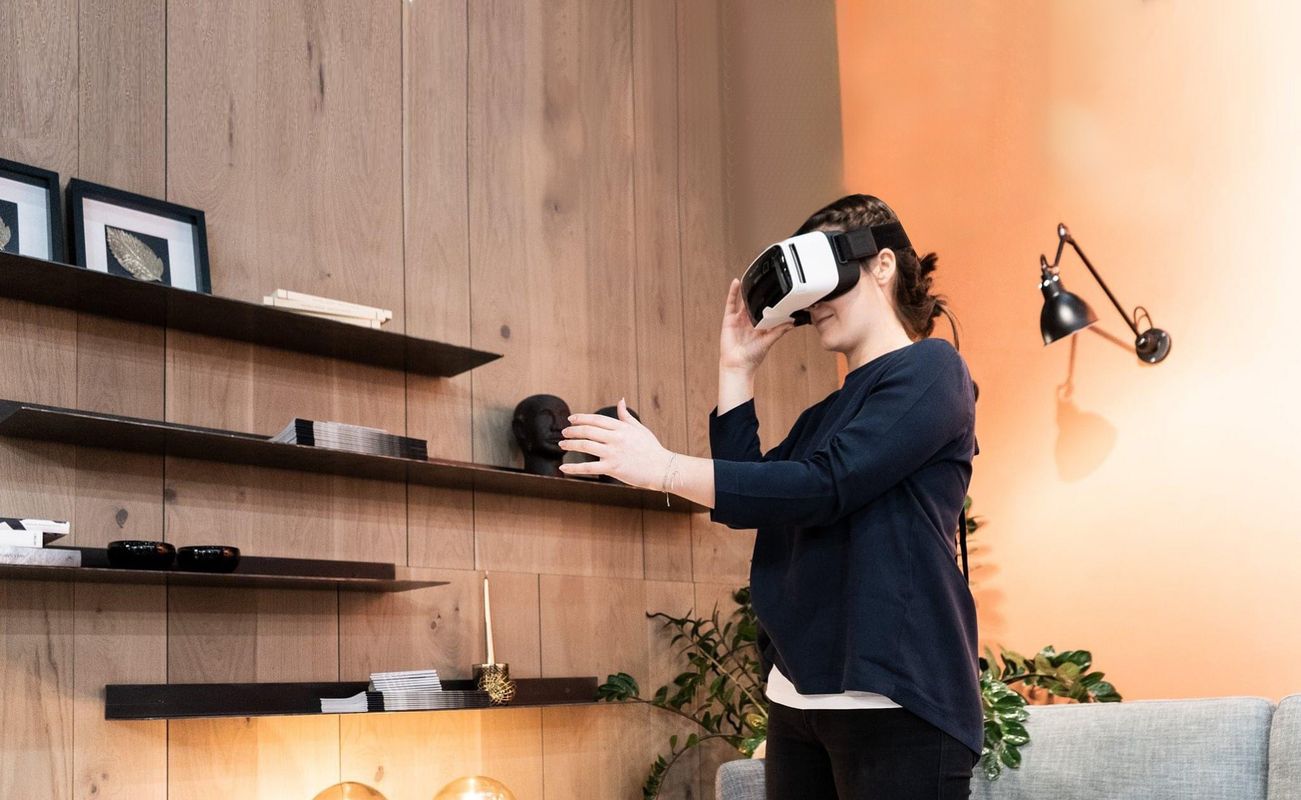 Eine Person trägt eine VR-Brille und steht in einem modernen Raum mit Holzregalen, Pflanzen und einer Couch.