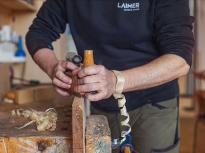 Ein Handwerker bearbeitet ein Holzstück mit einem Hobel. Holzspäne liegen auf der Werkbank.